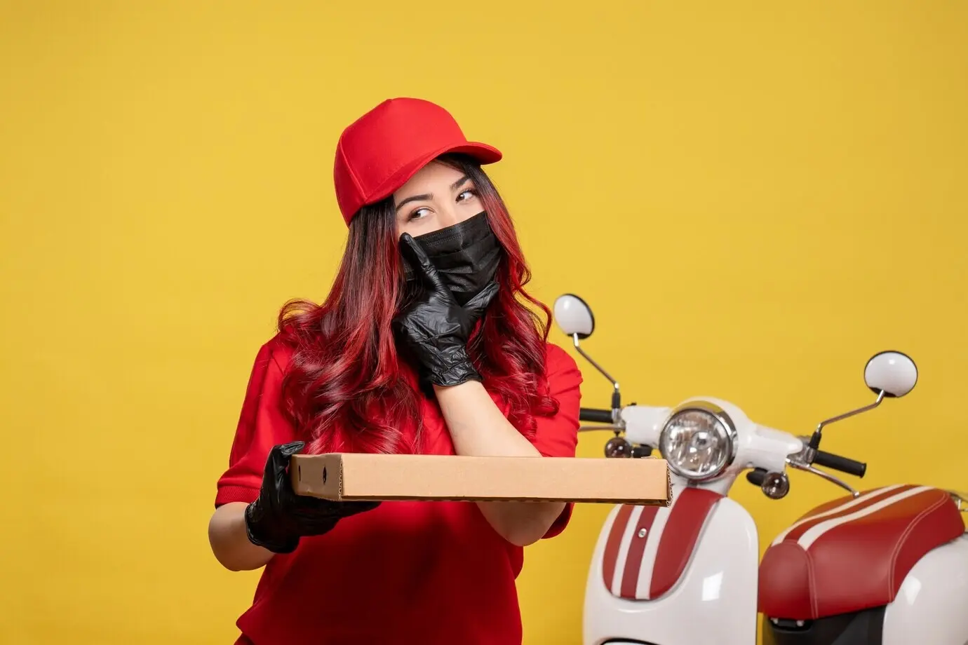Vorderansicht einer weiblichen Kurierin mit Maske und Essenslieferbox vor gelber Wand.